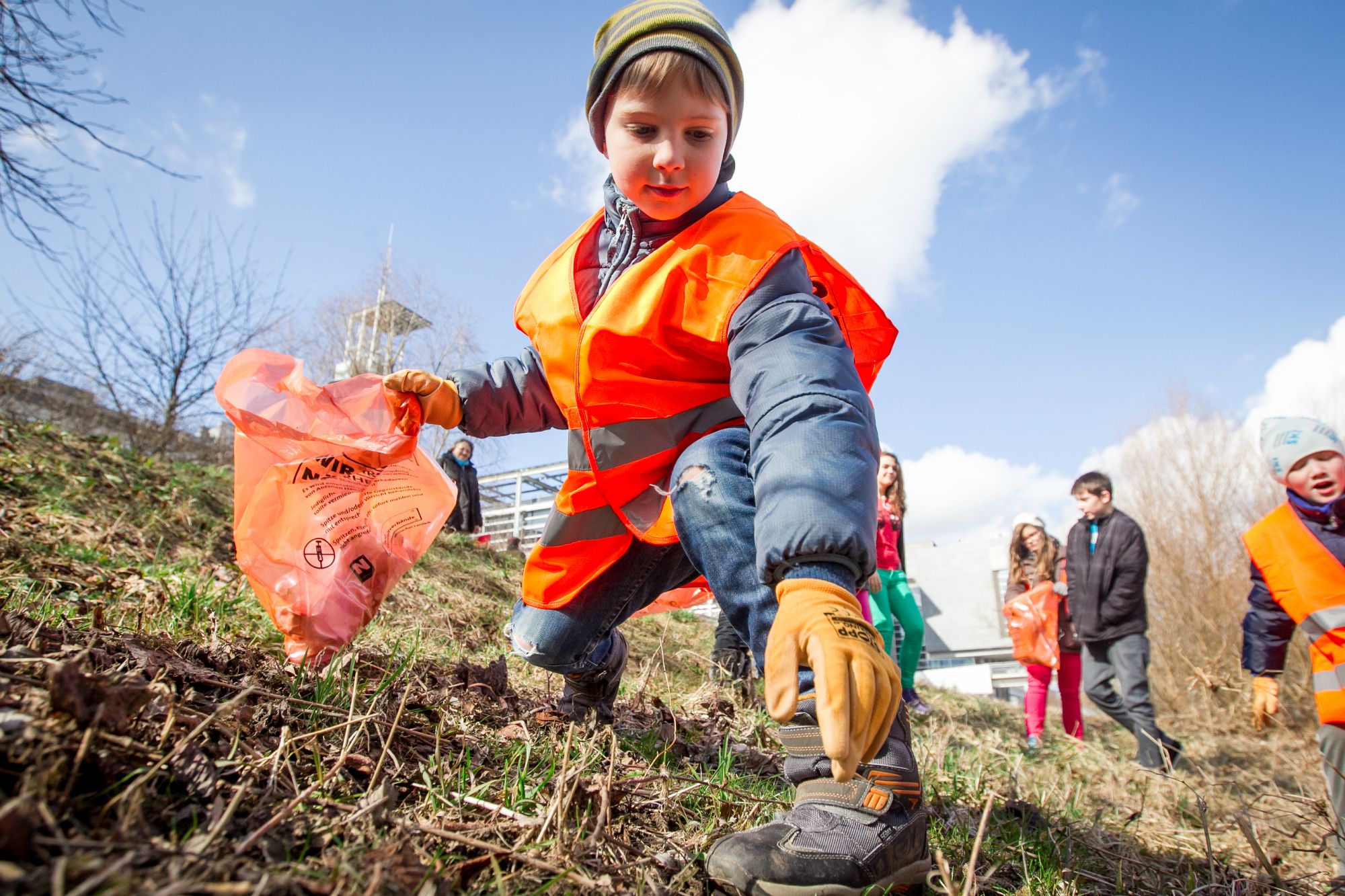 Zu sehen sind Kinder, die mit Warnwesten, Handschuhen und Sammelsäcken ausgestattet sind, Abfälle aus der Natur aufsammeln.